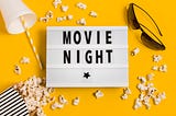 White clipboard with the words MOVIE NIGHT surrounded by popcorn, sunglasses and a drink cup. All on a bright yellow background