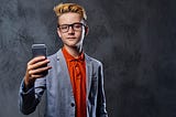 a smug-looking blond boy with glasses wearing a blazer holds up his cell phone while looking in to the camera