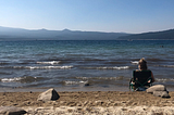 Girl sitting in a low profile camp chair along the shoreline of a lake with mountains in the distance