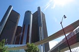 A photograph of the sun shining down over the GM Renaissance Center from the view of right under a People Mover track. One lamppost and two trees also add to its beauty. God bless and I love you!