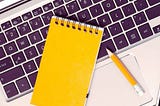 Otwarty laptop, ołówek, telefon i notes na klawiaturze. Obraz przedstawia temat — gdzie szukać pracy zdalnej?
