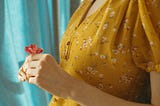 A cropped image of a woman wearing a mustard-colored, flowered dress holding a small red flower in her hands in front of her chest.