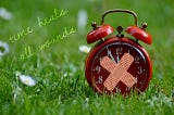 Image of an old-fashioned red alarm clock on the grass with two plasters stuck to it in a cross on the front, and to the left of it are the words: time heals all wounds.