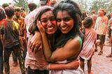 Two UC Berkeley students covered in colorful powder embrace and smile during the 2023 Holi Festival on campus.