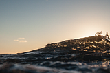 Photograph of a wave at sunset. The peak of the wave is highlighted by the sun.