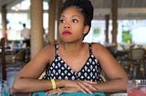 Black woman sitting at an empty dining space staring thoughtfully into the distance.