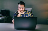 A man with glasses sitting in front of a laptop and looking concerned.