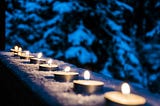 row of votive candles sitting outside on a handrail overlooking evergreens dusted with snow in the evening