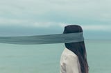 A woman standing by the ocean, a veil in front of her eyes