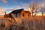 What looks like an old, rundown homestead house with aging wood siding and missing windows, in a field of grass next to a bare tree and against a partly cloudy but sunlit sky.