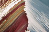 A landscape photograph of the Rainbow Mountain found in Peru. There’s a person standing three forths in the picture from bottom to top, gazing its beauty.