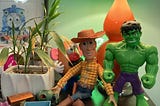 A photograph taken by author of author’s son’s Woody doll with his toy friends including Thomas and Friends, The Hulk, and My Potato Head next to bamboo plants.