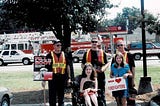 Two young women with three firefighters in front of fire truck.