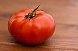 Photograph of one red, ripe tomato, on a brown wooden surface.