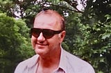 Photo of a man in sunglasses outside with a butterfly that landed on his bald head.