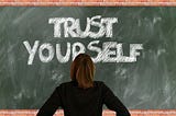 Blackboard with phrase, “Trust Yourself” written and person standing in front of it reading the text
