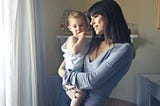 A woman holding a toddler and looking reflectively out the window.