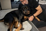 A man dressed in black and his Rottweiler pose for the camera. The dog is on a trampoline bed in a home office, and the man sits on the floor beside her. Her paw rests on his knee as a loving gesture of companionship. Both seem to be smiling.