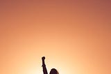 standing female figure with right fist raised in the air, silhouetted by a sunlight open sky