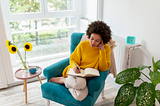 Black woman wearing yellow sweater and sitting on a teal sofa, writing in a notebook.