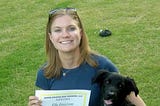 Meredith Emerson is grinning alongside her black dog, Ella, displaying a certificate for Ella’s graduation from a dog training program.