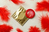 A gold foil condom wrapper lies on a light textured surface next to an unrolled red condom. Surrounding the items are several bright red feathers, adding a soft, vibrant contrast to the scene. The lighting is even, and the image has a clean, playful feel.