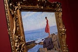 Close up of gold frame against a red wall, showing a painting of a woman in a red dress on a rock, looking out to sea