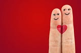 Two fingers close together, the shorter one has female eyes and mouth drawn on it, the taller has male eyes and a smile drawn on it, Each has half a red heart which — side by side — make a whole heart