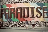 A picture of a couple holding hands in front of a graffiti wall with the word “paradise” in huge brown letters on a background of bright colors.