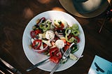 A colorful Greek salad on a white plate featuring fresh cucumbers, red bell peppers, red onions, black olives, and two portions of white cheese (likely feta or mozzarella) sprinkled with herbs and black pepper. A fork and knife rest on the plate. The meal is photographed from above on a wooden table with a straw hat, what appears to be a book or magazine with an eagle image, and other dining accessories visible around the edges of the frame.
