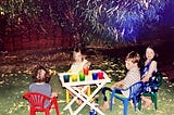 Four children sit on colourful chairs in a backyard at night. There is a table with snacks in front of them.