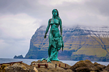 Photo of bronze sculpture of Selkie Seal-Woman standing on a rock in a bay in The Faroe Islands.