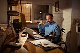 A man in a wheelchair sits at a desk in a home office smiling at his computer with a cat on his lap.