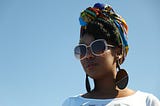 J LeShaé pictured from the shoulders up in shades and an African print head wrap against the sky. Photographer: Krystal René.