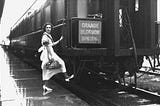 A 1930s black and white photo of a woman with her foot on the step on the last car of a train. Her hand is holding the handrail. She is wearing a white dress and white shoes and carrying a purse.