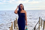 Picture of brown skinned black woman smiling. She is wearing a long black sleeveless dress and is standing on the dock with river behind her. The sky is clear with a few clouds.