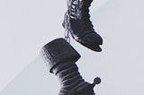 Two worn-out black boots hang from chains against a greyish background