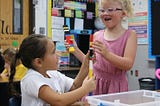 Two young girls, about age 7, one white with blonde hair and glasses, and one brown-skinned with dark hair in a pony tail, work together to build a tower using Lego bricks
