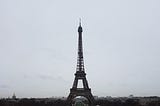 A photo of the Eiffel tower from a distance, set against a dim grey overcast sky.