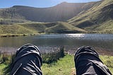 Man relaxed and looking over knees at a lake and hills in the sunshine.