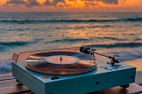 The image is of the shoreline of an ocean at sunset. In front of the water is a small record player sitting on a wooden table. On it is a blue record player with a vinyl album sitting on it.