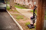 Image shows a curb between a street and tree-lined sidewalk. An abandoned upright vacuum rests there, awaiting garbage collection.