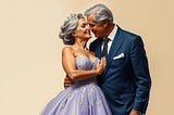Image shows an older man in a suit and an older woman in a ballgown and a tiara on her hair, hugging and looking into each other’s eyes.
