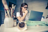 female looking thoughtfully at a laptop screen at her messy desk