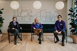 Kalda smiling team sat in a WeWork. From left: Al Dee, white person with short asymetric hair. Charlotte Fountaine, white woman with blonde hair. Daniel Botcherby, mixed race man with black hair and a purple. Kalda jumper.