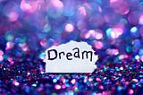 Purple, pink, and blue sparkling gems with a torn corner of the paper in the center. The word “dream” is written on the paper.