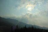 The sun glowing behind the clouds in a mountain scene.
