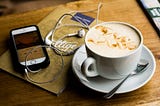 Phone with earbuds listening to a podcast episode with a cup of coffee in a coffee shop.