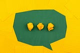 On a green table, pieces of yellow lined paper are arranged in an oval to create a speech bubble, which contains three balls of scrunched-up yellow paper to indicate conversation.