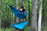 Kaitlyn sits in the top hammock of a stack of two, sipping coffee from an IBM branded mug and gripping the fabric with her free hand, as the dog and person in the lower hammock are being flipped out onto the forest floor. (Dog and human are fine and still attempting crazy stunts, today.)